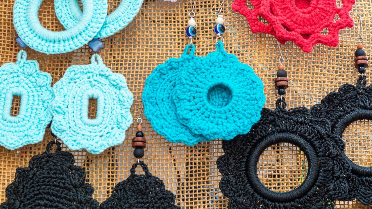 Imagem com diversos brincos de crochê com formatos arredondados em cores azul, preto e vermelho. Os brincos estão pendurados em tecido de juta.