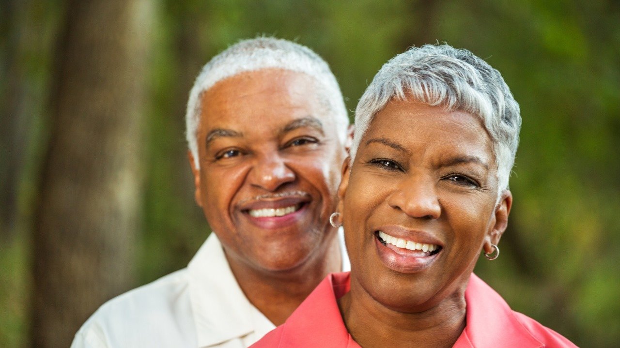 casal de pele negra, cabelos brancos e ambos sorriem para a foto de perfil. Ao fundo cor verde.