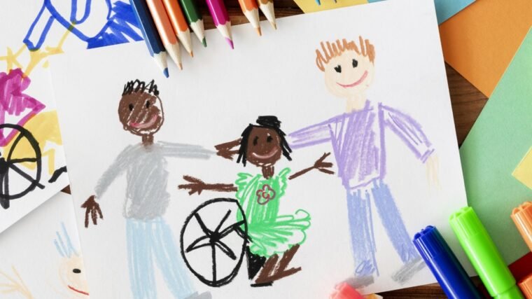 Imagem com uma folha de papel branca desenhada com 3 desenhos de uma menina de pele negra em uma cadeira de rodas e dois meninos em pé ao lado dela. Na parte superior tem diversos lápis de cor.