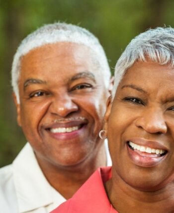 casal de pele negra, cabelos brancos e ambos sorriem para a foto de perfil. Ao fundo cor verde.