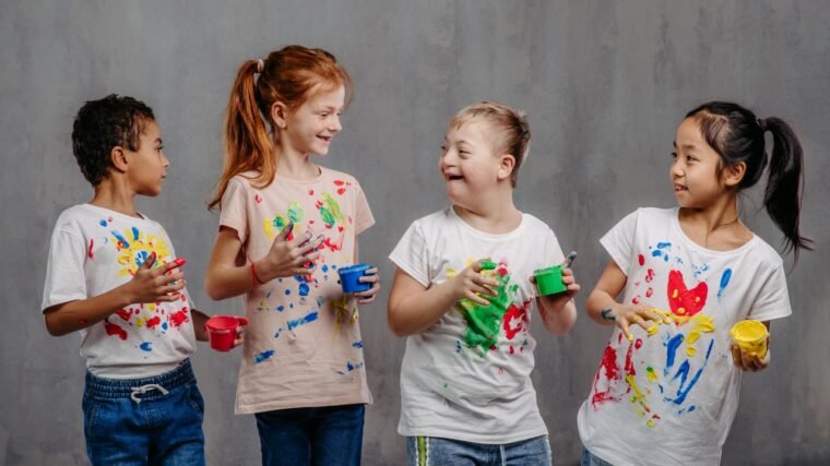 card co 3 crianças e todas estão sorrindo e vestem camisetas manchadas de tinta colorida. Elas estão com as mãos sujas de tinta. O fundo da imagem é opaca.