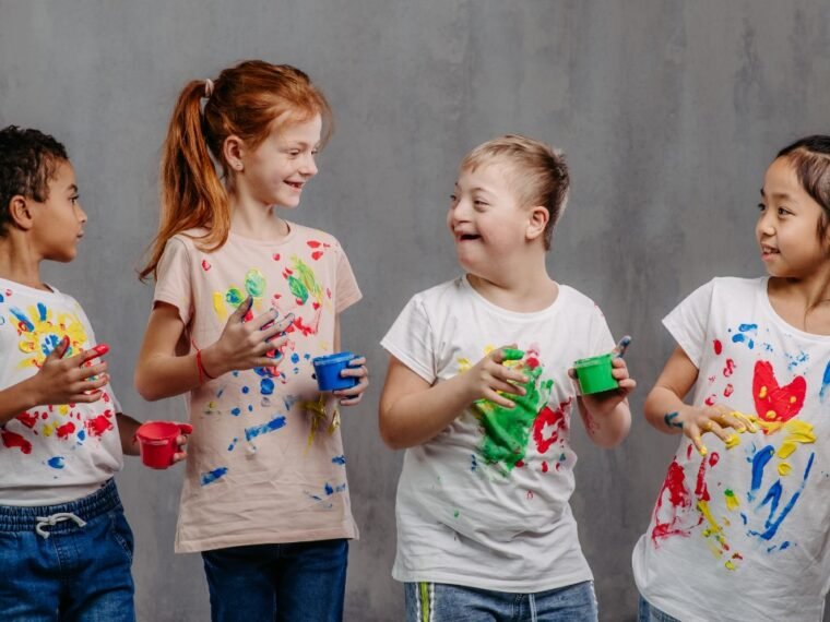 card co 3 crianças e todas estão sorrindo e vestem camisetas manchadas de tinta colorida. Elas estão com as mãos sujas de tinta. O fundo da imagem é opaca.