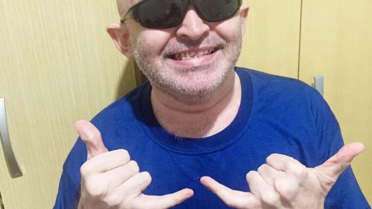 foto do Alex Garcia, homem de pele clara, careca, usa óculos pretos e veste camiseta azul. Ele está de perfil da cintura para cima e as duas mãos para cima com o polegar e o dedo mindinho para cima.
