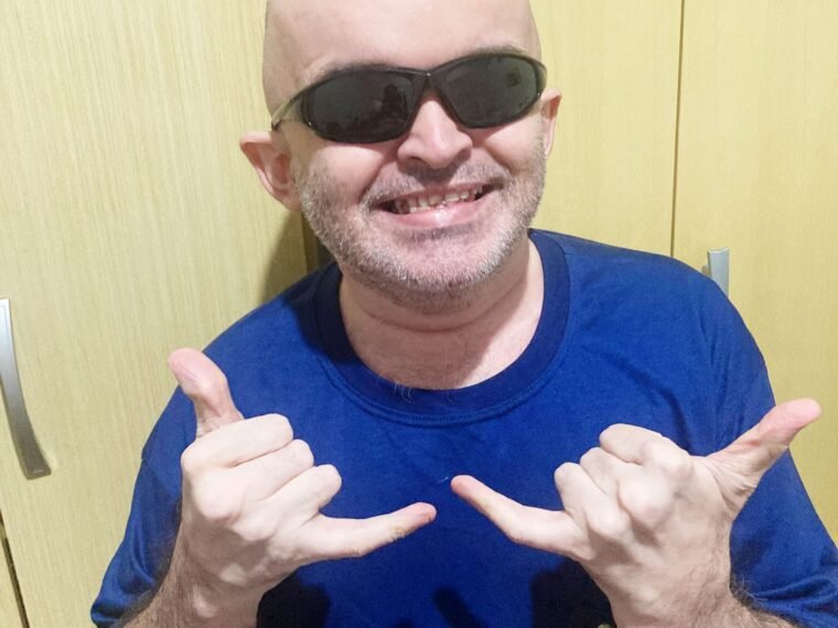 foto do Alex Garcia, homem de pele clara, careca, usa óculos pretos e veste camiseta azul. Ele está de perfil da cintura para cima e as duas mãos para cima com o polegar e o dedo mindinho para cima.