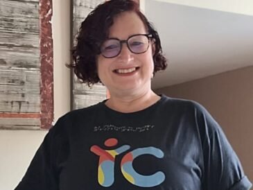 Mulher de pele clara, cabelos curtos, usa óculos de grau, vestida de blusa preta com o logo da Tudo Sobre Inclusão e calça verde. Ao fundo parede com 2 quadros e um holofote.