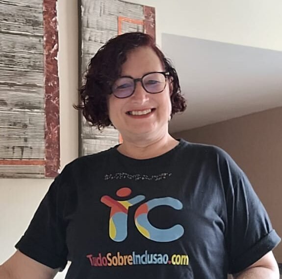 Mulher de pele clara, cabelos curtos, usa óculos de grau, vestida de blusa preta com o logo da Tudo Sobre Inclusão e calça verde. Ao fundo parede com 2 quadros e um holofote.