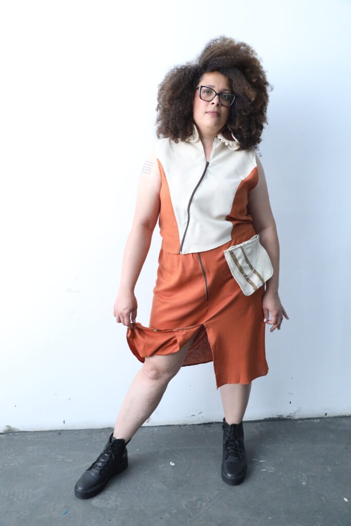 Modelo Ana Maria posa com o look três, ela segura com a mão direita a aba frontal da saia para o lado.