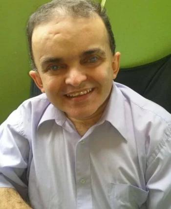 Imagem do Alex sentado, foto de perfil, homem de pele clara, rosto arredondado, olhos castanhos e camisa social de manda curta clara.