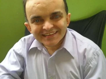 Imagem do Alex sentado, foto de perfil, homem de pele clara, rosto arredondado, olhos castanhos e camisa social de manda curta clara.