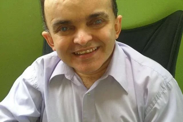 Imagem do Alex sentado, foto de perfil, homem de pele clara, rosto arredondado, olhos castanhos e camisa social de manda curta clara.