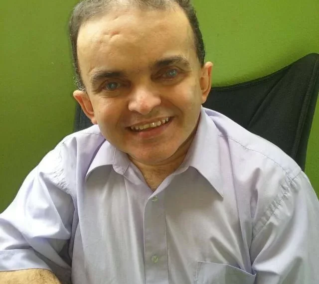 Imagem do Alex sentado, foto de perfil, homem de pele clara, rosto arredondado, olhos castanhos e camisa social de manda curta clara.